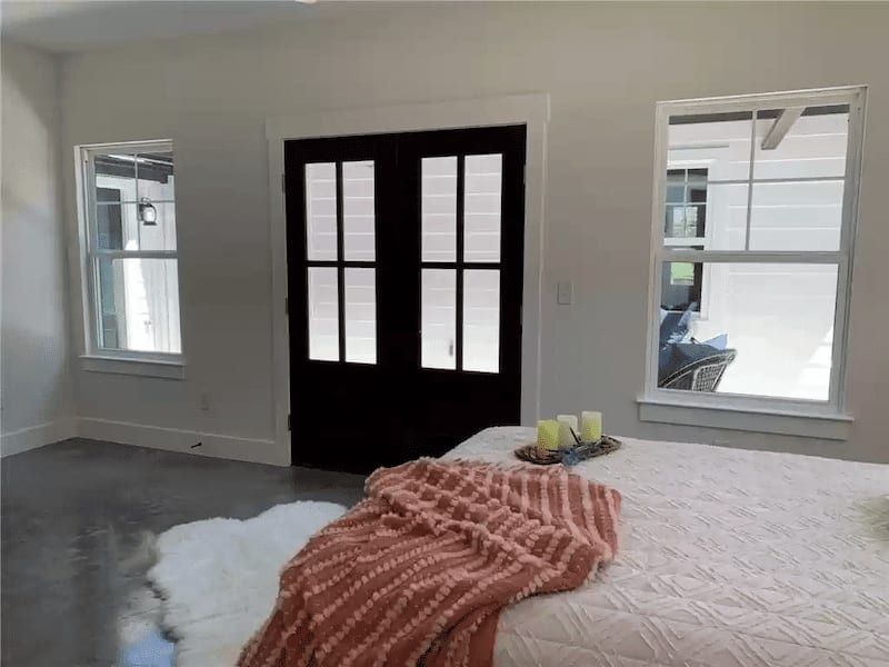 Bedroom with black French doors to a porch, polished concrete floor and a pair of white-trimmed windows