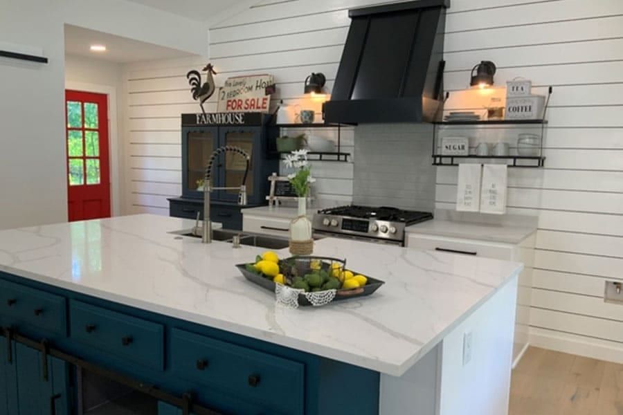 Kitchen with a teal island, white marble counters, a black range hood and shiplap walls