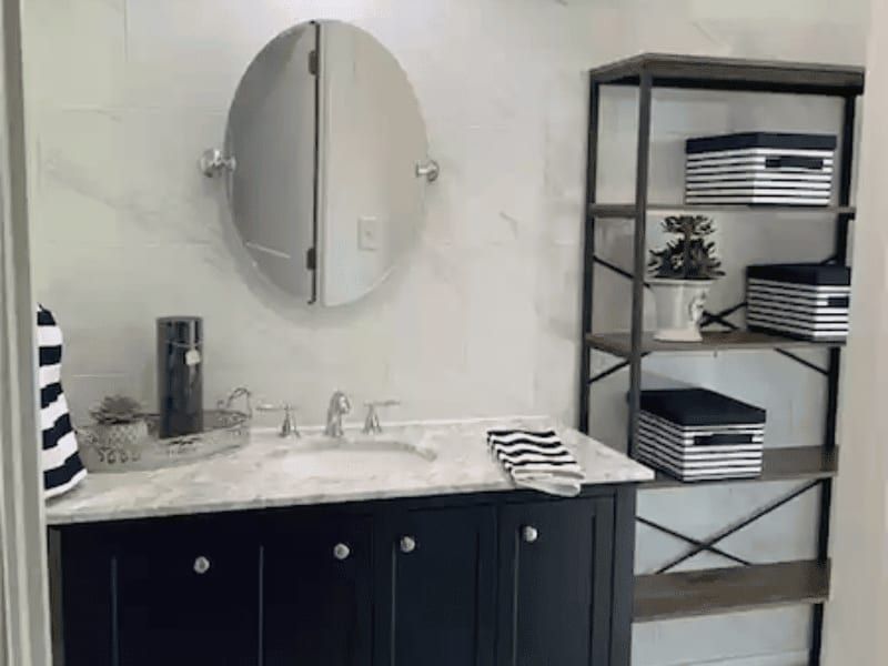 Bathroom vanity in navy cabinetry with a marble top, an oval pivot mirror and an open metal shelf beside it