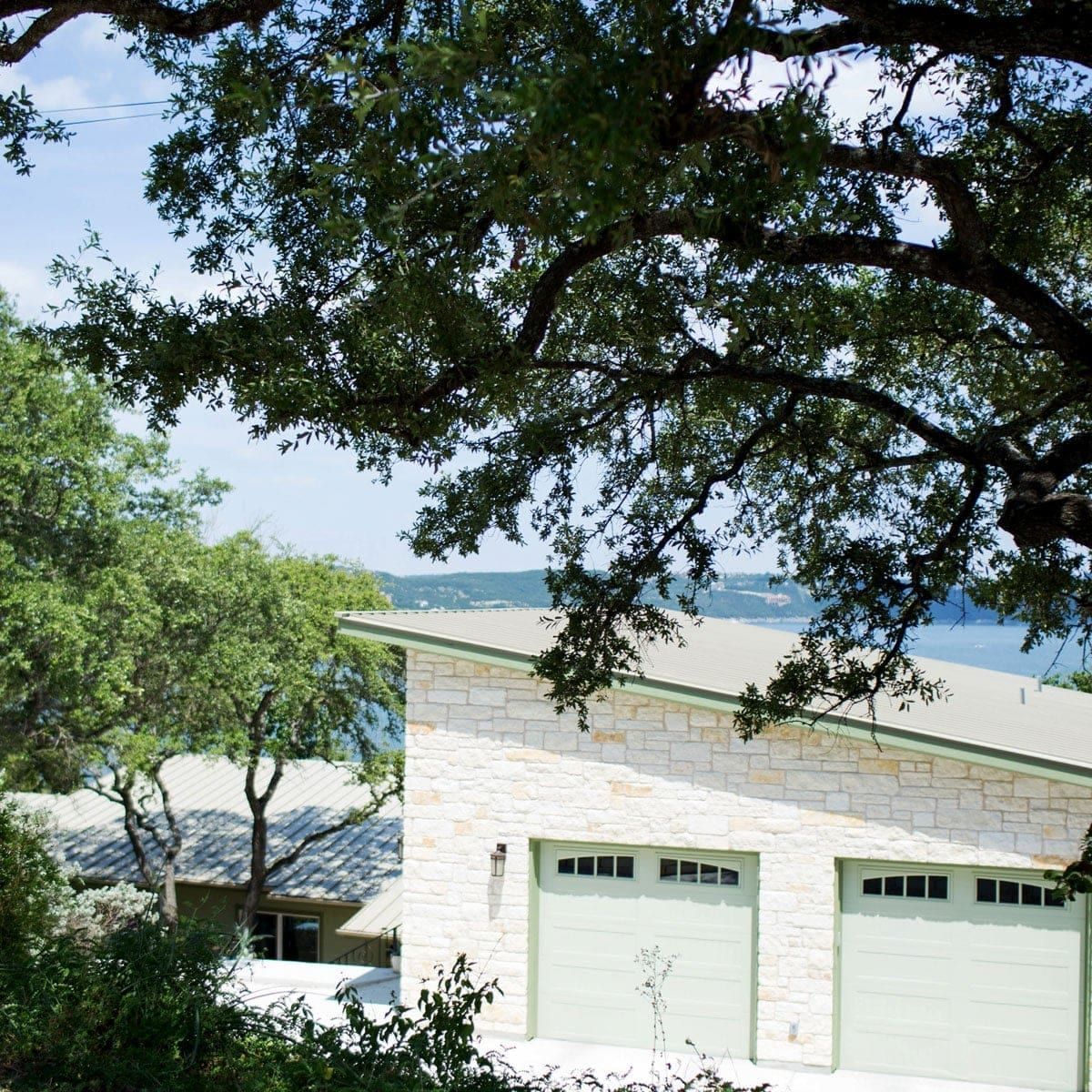 Stone garage wing with a standing seam metal roof, framed by live oaks with Lake Travis beyond