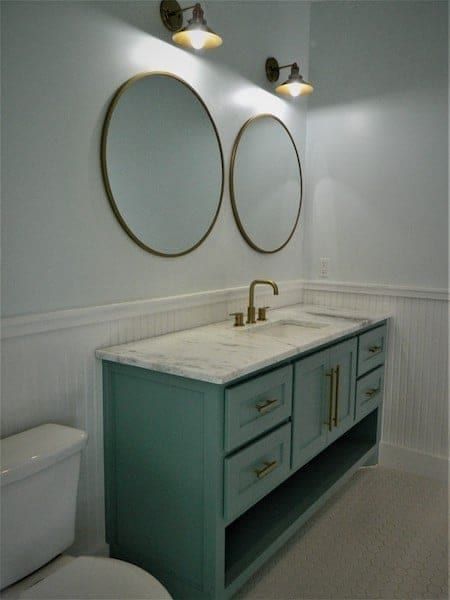 Bathroom with a green vanity, a marble top, two round brass-framed mirrors and beadboard wainscot